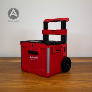 Può includere: Cassetta portautensili rossa e nera con maniglia retrattile e ruote. La cassetta degli attrezzi ha il logo "Milwaukee" sul lato. L'immagine mostra anche il logo "Apex Makes" nell'angolo in alto a sinistra.