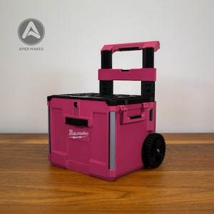 Puede incluir: Un carro de herramientas rosa con asa y ruedas negras. El carro tiene el texto "Make-maker" en la parte delantera. Está sobre una superficie de madera.