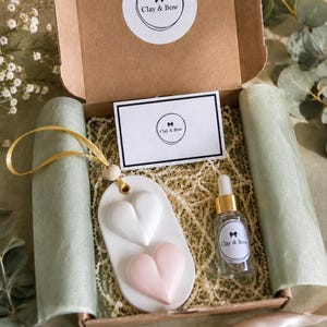 May include: A brown cardboard gift box containing a white oval clay ornament with one white and one pink heart, a gold ribbon, a small glass dropper bottle with a gold cap, and a white card, all labelled with the Clay & Bow logo.