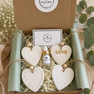 May include: A brown cardboard gift box containing four white heart-shaped clay ornaments with various embossed patterns, including one with the word LOVE in gold. A small glass dropper bottle with a Clay & Bow label and a branded card are included.