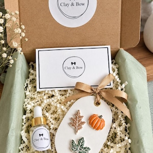 May include: A cardboard gift box containing a white oval clay ornament with a brown leaf, an orange pumpkin, and a green leaf. A gold ribbon is tied to the ornament. A small glass dropper bottle and a white card both feature the Clay & Bow logo.