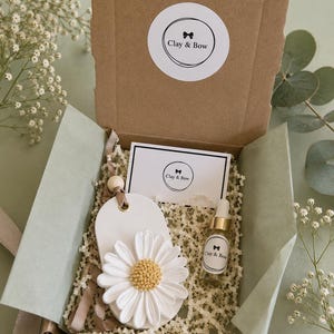 May include: A brown cardboard gift box containing a white oval clay ornament with a sculpted white daisy featuring a gold centre, a small glass dropper bottle with a Clay & Bow label, and a rectangular card with the Clay & Bow logo.