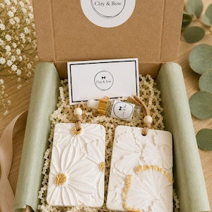 May include: A brown cardboard gift box containing two rectangular white clay air fresheners with embossed floral and leaf patterns, accented with gold paint. Includes a small glass dropper bottle with a gold cap and a white card labeled Clay & Bow.