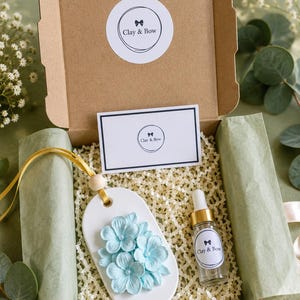May include: A brown cardboard gift box containing a white oval ceramic diffuser with light blue floral accents, a small glass dropper bottle, and a rectangular card. The box lid and items feature a circular logo with the text Clay & Bow.