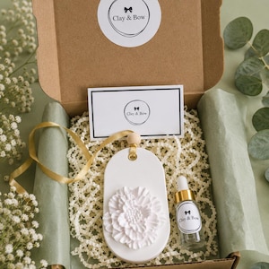 May include: A brown cardboard gift box containing a white oval-shaped clay ornament with a raised flower design, a small glass dropper bottle with a gold cap, and a white card. Both the box lid and card feature the text Clay & Bow.