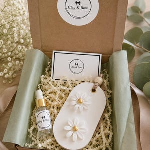 May include: A brown cardboard gift box containing a white oval-shaped clay ornament with two white daisy flowers featuring gold centres, a small glass dropper bottle with a gold cap, and a white card, all labelled with the Clay & Bow logo.
