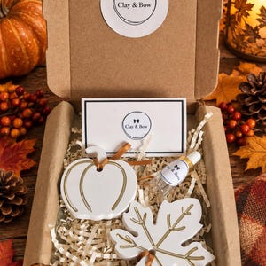 May include: A brown cardboard gift box containing a white ceramic pumpkin ornament and a white ceramic maple leaf ornament, both with gold-coloured accents. A small glass vial with a Clay & Bow label and a branded card are included in the box.