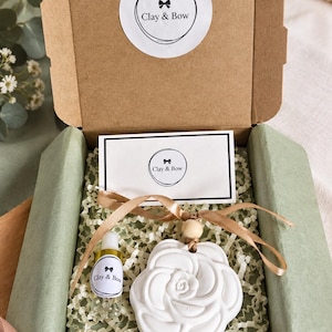 May include: A brown cardboard gift box containing a white rose-shaped clay ornament with a wooden bead and gold ribbon, a small clear glass bottle with a Clay & Bow label, and a white card with the Clay & Bow logo, all nestled in green paper.