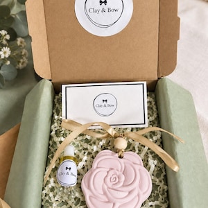 May include: A brown cardboard gift box containing a light pink rose-shaped clay ornament with a gold ribbon and a small clear glass bottle of oil. The box features a white sticker and a card both printed with the text Clay & Bow.
