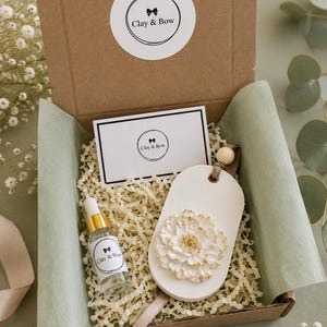 May include: A brown cardboard gift box containing a white oval clay ornament with a raised white and gold flower, a small glass dropper bottle with a Clay & Bow label, and a rectangular card with the Clay & Bow logo, all nestled in paper filler.