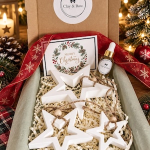 May include: A cardboard gift box containing three white star-shaped ornaments with jute twine and wooden beads, a small glass dropper bottle labelled Clay & Bow, and a rectangular card with a wreath graphic and the text Merry Christmas.