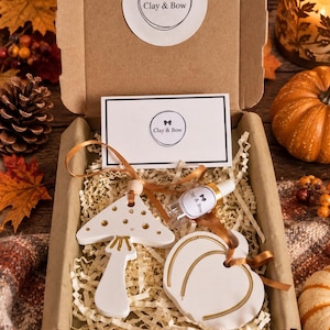May include: A cardboard gift box containing a white clay mushroom ornament with gold accents, a white clay pumpkin ornament with gold trim, a small glass dropper bottle labelled Clay & Bow, and a rectangular card with the Clay & Bow logo.