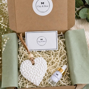 May include: A brown cardboard gift box containing a white textured heart-shaped ornament with a wooden bead and brown ribbon, a small glass dropper bottle with a gold cap, and a white card, all labelled with the Clay & Bow logo.