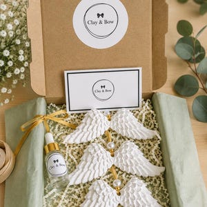 May include: A brown cardboard gift box containing three white textured angel wing ornaments strung on a gold cord with white beads. A small glass dropper bottle with a Clay & Bow label, a branded card, and a circular sticker are included.