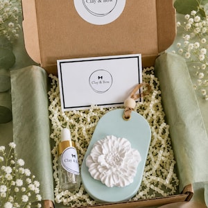 May include: A gift box containing a light blue oval clay ornament with a white floral relief, a small glass dropper bottle with a gold cap, and a rectangular card. Both the card and the bottle feature a circular logo with the text Clay & Bow.