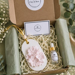 May include: A white oval hanging ornament featuring a cluster of light pink sculpted flowers, suspended by a gold ribbon and a wooden bead. The set includes a small glass dropper bottle with a gold cap, both labelled Clay & Bow, inside a box.