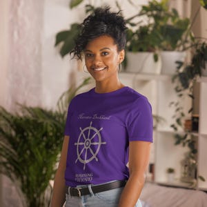 May include: A purple t-shirt featuring a silver ship's wheel design. The shirt has the text "Mercator Deckhand" above the wheel and "Bloodsong of Wygaro" below. The model is wearing a black belt and blue jeans.