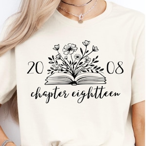 Puede incluir: Camiseta color crema con un diseño floral negro sobre un libro abierto. Los números "20" y "08" flanquean el arreglo floral, con las palabras "chapter eighteen" debajo en cursiva.