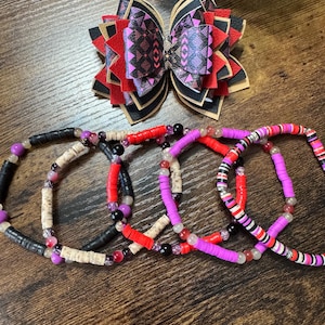May include: A handmade hair bow with a geometric pattern in red, black, and purple, and several beaded bracelets in various colors. The bracelets feature black, red, purple, and white beads, with some bracelets having a mix of colors.