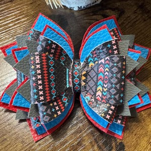 May include: A handmade hair bow with a layered design. The bow features a black base with colorful geometric patterns, accented by red, blue, and gray layers. The bow is likely made of faux leather or a similar material.