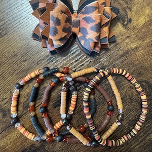 Puede incluir: Un lazo para el cabello hecho a mano con un diseño de estampado de leopardo y varias pulseras con cuentas. El lazo presenta capas de cuero sintético marrón, negro y con estampado de leopardo. Las pulseras están hechas con cuentas coloridas en tonos marrón, naranja y negro.