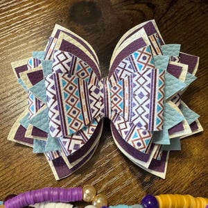 May include: A large, layered hair bow with a geometric pattern in shades of purple, blue, and cream. The bow features a central design with vertical stripes and diamond shapes. A beaded necklace with various colors and shapes is in the foreground.