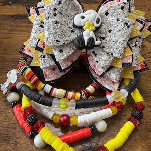 May include: A multi-layered hair bow featuring silver glitter, white, and yellow accents with a Snoopy character centerpiece. Attached are several beaded bracelets made of black, white, red, and yellow clay discs, round beads, and a small Snoopy charm.