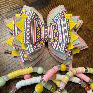 May include: A handmade hair bow with a geometric pattern in shades of pink, yellow, green, and white. Several beaded bracelets in pastel colors are also visible, featuring a mix of beads and disc-shaped elements.