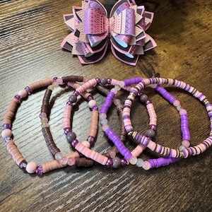 May include: A collection of beaded bracelets in shades of pink, brown, and purple, with a matching decorative bow. The bracelets feature disc-shaped beads and small round beads. The bow is made of layered paper with a patterned design.