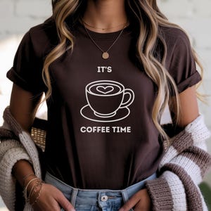May include: A dark brown t-shirt with the text "IT'S COFFEE TIME" and a white graphic of a coffee cup with a heart-shaped design in the foam. The shirt has rolled-up sleeves.