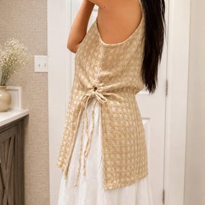 May include: A sleeveless beige and white checkered tunic featuring a side drawstring closure with thin fabric ties. The tunic is layered over a long white skirt. The checkered pattern consists of a grid design in light tan and white tones.