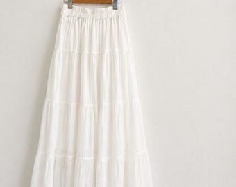 May include: A floor-length white skirt featuring a tiered design with a gathered elastic waistband. The skirt is made of a lightweight, textured fabric and hangs from a wooden hanger, showcasing a full, flowing silhouette with multiple horizontal seams.