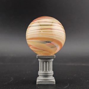 May include: A decorative glass sphere with orange, tan, and white horizontal stripes, resting on a gray, miniature column. The sphere has a marbled appearance. The column has a square base and a fluted design.
