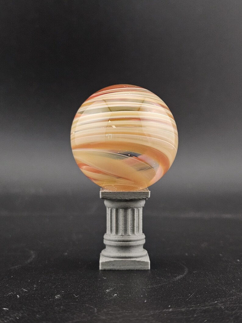 May include: A decorative glass sphere with orange, tan, and white horizontal stripes, resting on a gray, miniature column. The sphere has a marbled appearance. The column has a square base and a fluted design.