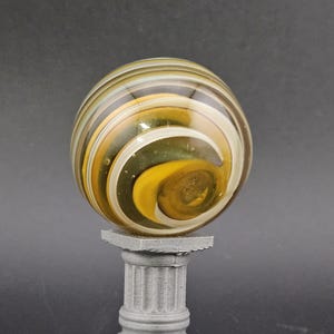 May include: A decorative glass marble with yellow, white, and brown swirls, resting on a small, gray, classical-style pedestal. The marble has a spiral design and is the focal point of the image.