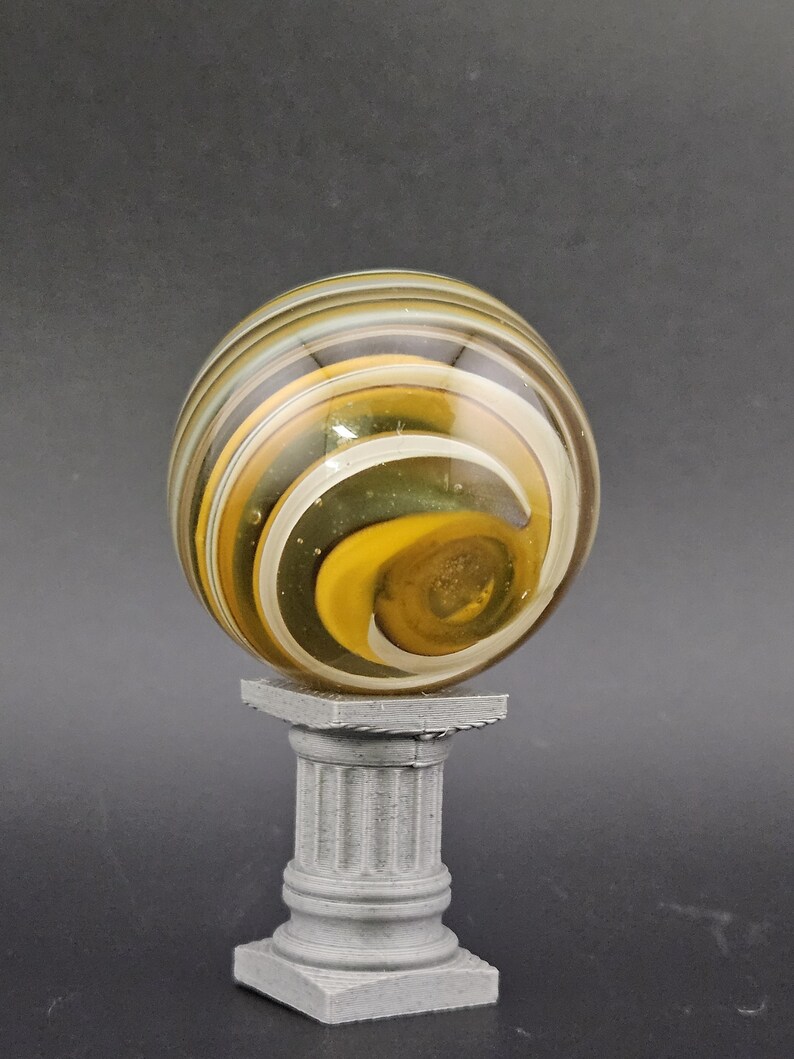 May include: A decorative glass marble with yellow, white, and brown swirls, resting on a small, gray, classical-style pedestal. The marble has a spiral design and is the focal point of the image.