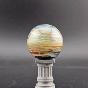 May include: A decorative glass orb with swirling colors of blue, yellow, and orange, resting on a small, gray, fluted column. The orb appears to be made of glass and has a polished, reflective surface. The column is square at the base and top.