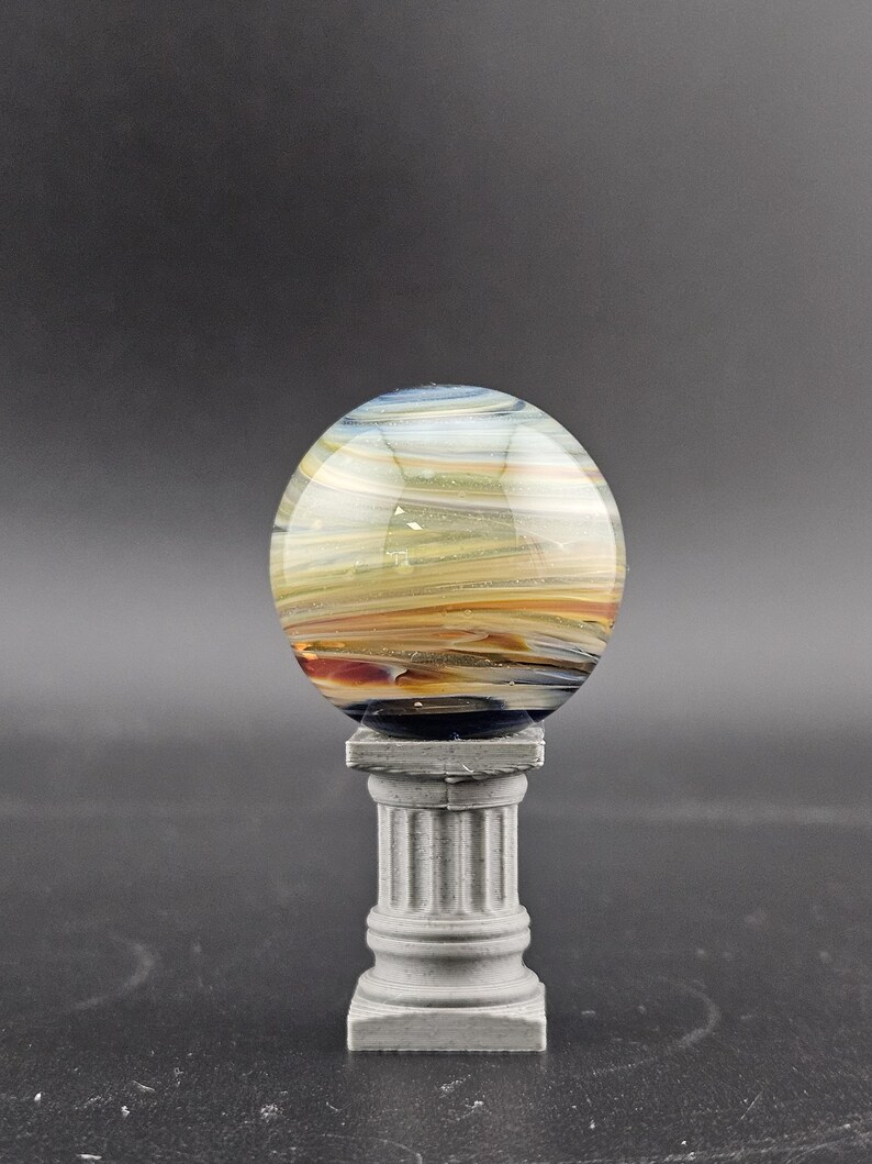 May include: A decorative glass orb with swirling colors of blue, yellow, and orange, resting on a small, gray, fluted column. The orb appears to be made of glass and has a polished, reflective surface. The column is square at the base and top.