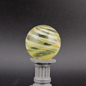 May include: A decorative glass sphere with yellow and white swirls, resting on a gray, fluted column pedestal. The sphere has a translucent quality, and the pedestal has a square base.
