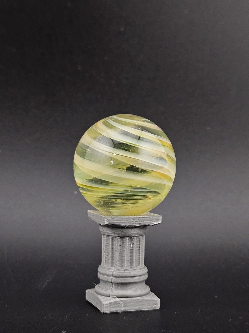 May include: A decorative glass sphere with yellow and white swirls, resting on a gray, fluted column pedestal. The sphere has a translucent quality, and the pedestal has a square base.