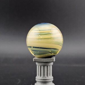 May include: A decorative glass sphere with yellow, blue, and green swirls, resting on a small, gray, classical-style pedestal. The sphere has a translucent quality, with visible internal patterns and bubbles. The pedestal is a miniature column.