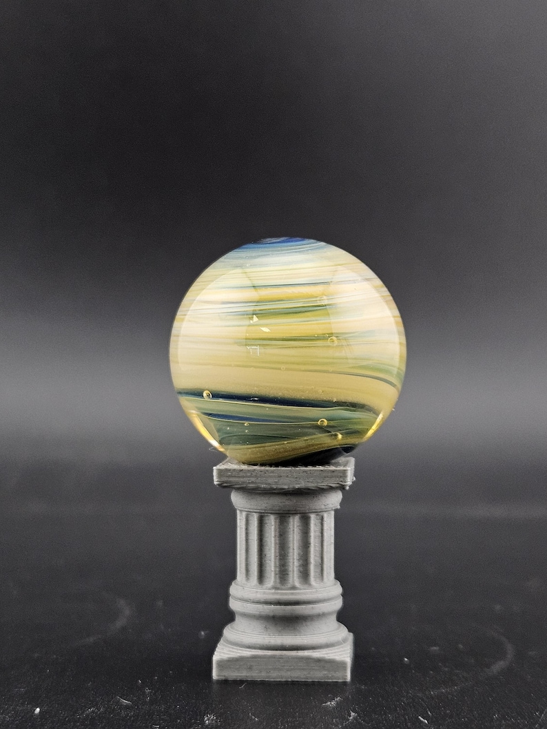 May include: A decorative glass sphere with yellow, blue, and green swirls, resting on a small, gray, classical-style pedestal. The sphere has a translucent quality, with visible internal patterns and bubbles. The pedestal is a miniature column.
