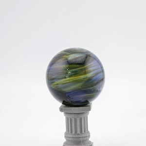 May include: A decorative glass sphere with swirling blue, green, and yellow colors, resting on a small, gray, classical-style column. The sphere appears to be made of glass and has a glossy finish.