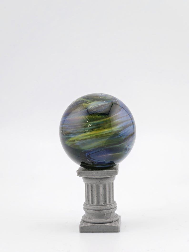 May include: A decorative glass sphere with swirling blue, green, and yellow colors, resting on a small, gray, classical-style column. The sphere appears to be made of glass and has a glossy finish.