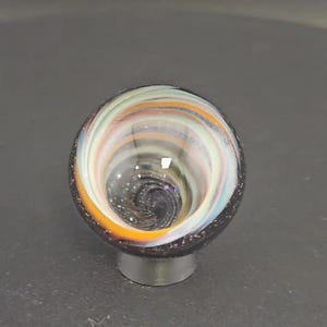 May include: A spherical glass object featuring a swirling pattern of white, orange, and light green colors. The center of the sphere displays a deep, dark vortex with subtle glitter-like flecks, while the outer rim is a solid dark charcoal color.