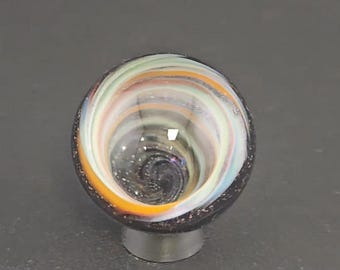 May include: A spherical glass object featuring a swirling pattern of white, orange, and light green colors. The center of the sphere displays a deep, dark vortex with subtle glitter-like flecks, while the outer rim is a solid dark charcoal color.