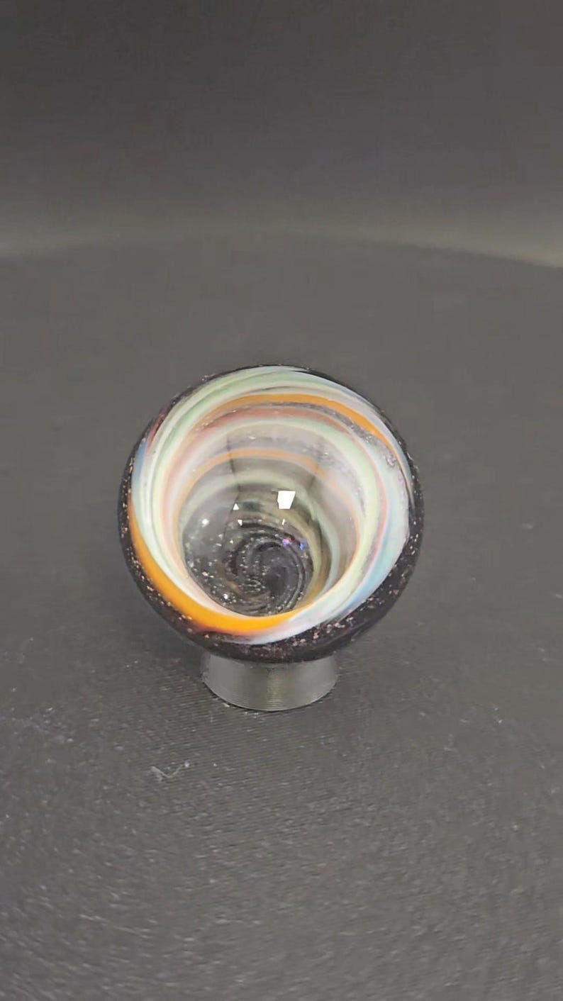 May include: A spherical glass object featuring a swirling pattern of white, orange, and light green colors. The center of the sphere displays a deep, dark vortex with subtle glitter-like flecks, while the outer rim is a solid dark charcoal color.