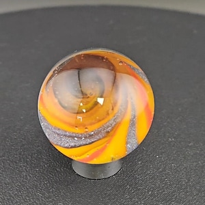 May include: A spherical glass marble featuring a swirling pattern of vibrant orange, yellow, and dark grey colours. The surface is smooth and transparent, revealing internal layers of colour and small air bubbles trapped within the clear glass sphere.