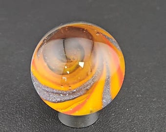 May include: A spherical glass marble featuring a swirling pattern of vibrant orange, yellow, and dark gray colors. The surface is smooth and transparent, revealing internal layers of color and small air bubbles trapped within the clear glass sphere.