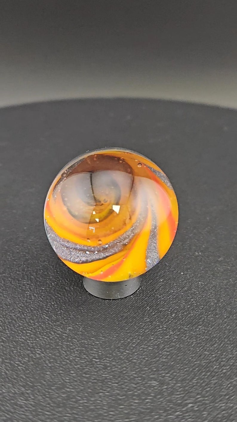 K&ouml;nnte beinhalten: Eine kugelf&ouml;rmige Glasmurmel mit einem wirbelnden Muster aus leuchtendem Orange, Gelb und Dunkelgrau. Die Oberfl&auml;che ist glatt und transparent und gibt den Blick auf innere Farbschichten und kleine Luftblasen im Inneren der Glaskugel frei.